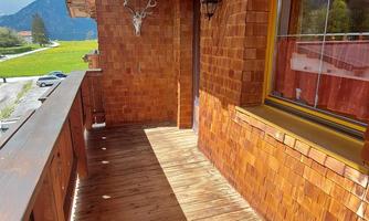 A cozy wooden balcony with a sunny wooden floor. In the background, green meadows and mountains are visible.