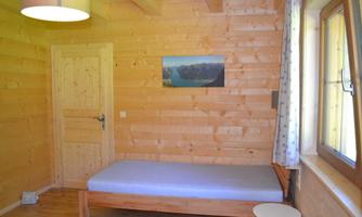 A simple room with wooden walls and a single bed. A picture of a landscape hangs on the wall, and there is a window with curtains.