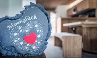 A heart-shaped cushion with the print "Alpenglück" and a red heart. In the background, a cozy apartment with wooden details can be seen.