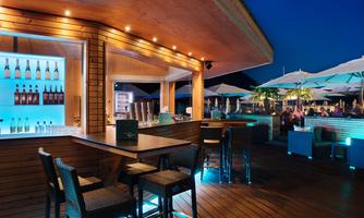 A stylish bar with wooden decor and blue lights at night. The terrace is equipped with sun umbrellas and tables, surrounded by guests.