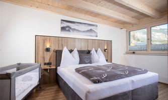 A modern bedroom with a large bed and wooden ceiling. A crib is next to the bed, and large windows provide plenty of daylight.