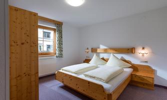A simple bedroom with a large wooden double bed and two pillows. The room has a window with curtains and a bright ceiling lamp.