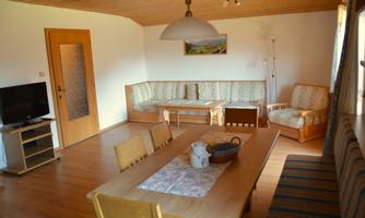 A cozy living room with a table and wooden chairs. In the room, there is a TV and a sofa set with a warm wooden floor and window light.