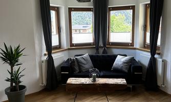 A cozy living room with a black sofa and a wooden table. Large windows provide plenty of daylight and a green view.