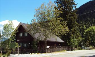 A traditional wooden house with a steep roof stands on a quiet street. In the background, trees and a hill can be seen.