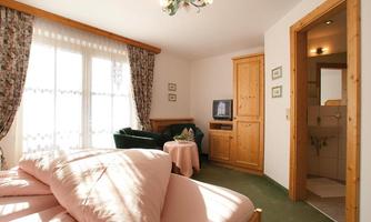 A cozy room with a double bed and pink blankets. There is a small table, a television, and a view of a bathroom.