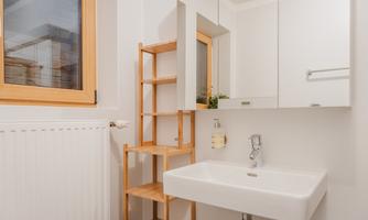 A modern bathroom with a white sink and a wooden shelf. Bright walls and a window create a friendly atmosphere.