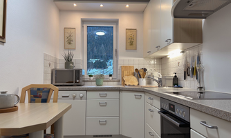 A modern kitchen with a window through which one can see the snowy landscape. There is a dining area, kitchen appliances, and light-colored furniture.