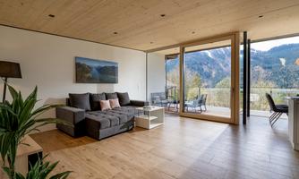 A modern living room with a cozy couch and large windows. The view extends to a beautiful mountain landscape.