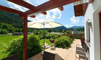 A sunny terrace with a sun umbrella and a grill. In the background, green meadows and mountains are visible.