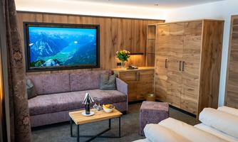 A cozy living room with a sofa, a table, and a large television. The room has a modern wood look and a pleasant atmosphere.
