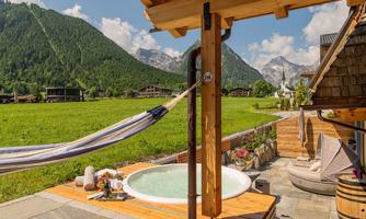 A relaxing outdoor area with a hot tub and a hammock. In the background, green meadows and picturesque mountains are visible.