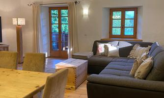 A cozy living room with a gray sofa and a wooden table. Large windows provide plenty of daylight and a view outside.