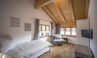 A cozy room with a wooden ceiling and large windows. The furnishings consist of a single bed, an armchair, and a television.