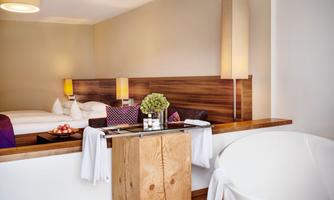 A modern hotel room with a large bed and stylish lamps. In the foreground, there is a freestanding bathtub and a wooden table with decorations.