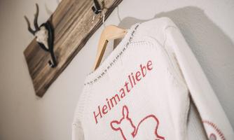 A knitted sweater with the print "Heimatliebe" hangs on a wooden hanger. In the background, antlers are mounted on a wooden wall.