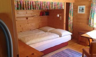 A cozy room with a double bed, wooden paneling, and colorful curtains. There is a table and a carpet that give the room a warm atmosphere.