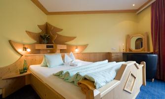 A cozy bedroom with a large double bed and wood-trimmed elements. The walls are painted yellow, and there is a mirror as well as an armchair.