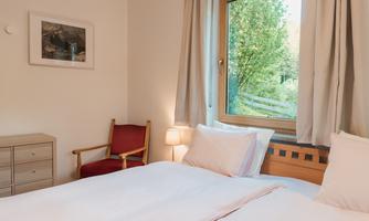 A cozy bedroom with two beds and a window through which plenty of daylight streams. In the room, there is a chair and a small lamp that create an inviting atmosphere.