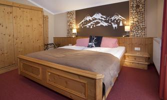 A cozy bedroom with a large bed and wooden furniture. On the wall, stylized mountains and warm lighting can be seen.