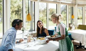 A restaurant with large windows and bright light. A server brings food to a couple sitting at the table.
