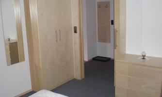 A simple bedroom with a wardrobe and dresser made of light wood. The room has a neutral carpet and good lighting.