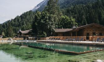 A quiet outdoor pool with clear water, surrounded by mountains and trees. In the background, wooden buildings are visible, creating a cozy atmosphere.