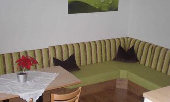 A cozy living area with a green corner sofa and a small table. On the table, there is a plant decoration, and there are cushions in dark brown.