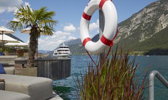 A relaxed lakeside view with a lifebuoy and modern furniture by the water. In the background, a ship and the mountains are clearly visible.