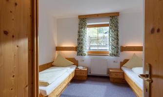 A cozy room with two single beds and wooden paneling. Daylight streams in through the window and there is a friendly atmosphere.