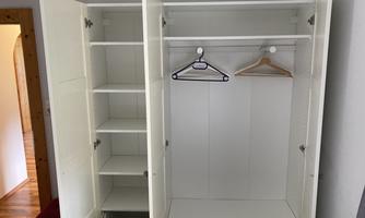 An open white wardrobe with several shelves and a hanger. The wardrobe has two doors and is located in a room with wooden flooring.