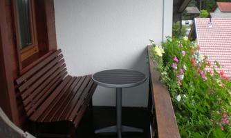 A cozy balcony with a wooden bench and a round table. Colorful flowers grow along the balustrade.