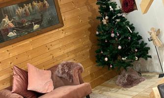 A cozy living room with a pink armchair and a Christmas tree. The walls are made of wood, and a painting hangs above the armchair.