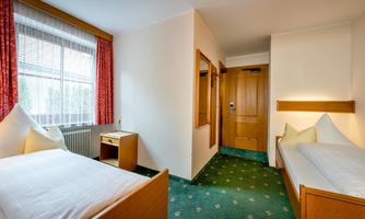A cozy room with two single beds, a desk, and a window. The walls are light, and the carpet is green with a pattern.