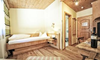 A cozy room with wooden decor, a single bed, and a small table. The room has natural light sources and an inviting view of the door to the bathroom.