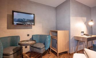 A modern room with two blue armchairs and a small table. A television is hung on the wall, and there is a wooden desk in the corner.