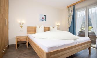 A modern bedroom with a large wooden bed and two pillows. Next to the bed is a nightstand and there are large windows with curtains.