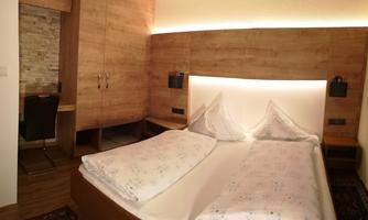 A modern bedroom with a double bed and wood paneling. Behind the bed there is a desk and in the background, a wardrobe.