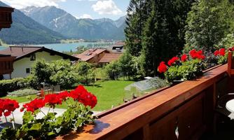 A beautiful view of a green landscape with a lake and mountains in the background. Red geraniums are blooming on the balcony.