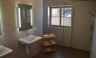 A modern bathroom with a large shower area and a bright, neutral color scheme. There is a sink, a mirror with LED lighting, and a towel cart.