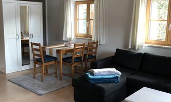 A cozy living space with a black sofa, a dining table, and chairs. The bright windows create a friendly atmosphere.