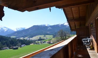 A terrace with a wooden railing and a view of green meadows and mountains. In the background, snow-capped peaks and a clear blue sky can be seen.