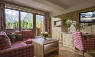 A cozy living space with a modern wooden interior and large windows offering a magnificent view of the mountains. The seating furniture is designed in a checkered pattern, and a flat-screen TV is mounted on the wall.