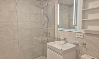 A modern bathroom with an elegant shower and a sink. The walls are tiled in a light, neutral tone.