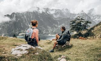 A beautiful mountain landscape with two hikers sitting on a rock and enjoying the view. In the background, impressive mountains and a clear lake can be seen.