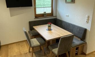 A cozy dining area with a wooden table and gray chairs. In the background, there is a window with a view and a small TV.