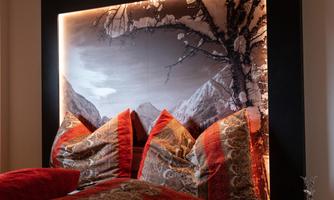A cozy bedroom with a large, illuminated headboard and colorful pillows. The rustic style is emphasized by the wooden ceiling and warm colors.
