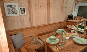 A cozy dining corner made of wood with a table and bench. The table is lovingly set with plates, glasses, and fresh fruits.