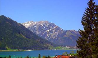 A picturesque landscape with snow-covered mountains and a clear blue lake. In the foreground, typical alpine houses and forest are visible.