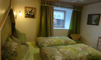 A cozy bedroom with two single beds and green bedspreads. The room has a large window with curtains and decorative pictures on the wall.